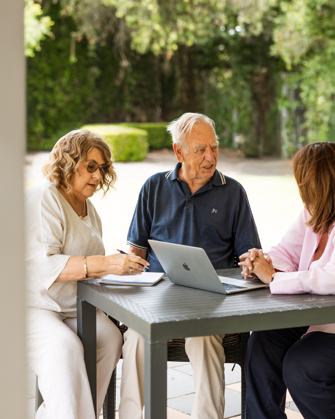 An older couple sit outdoors at a table with a laptop and notebook, discussing aged care fees with an advisor.