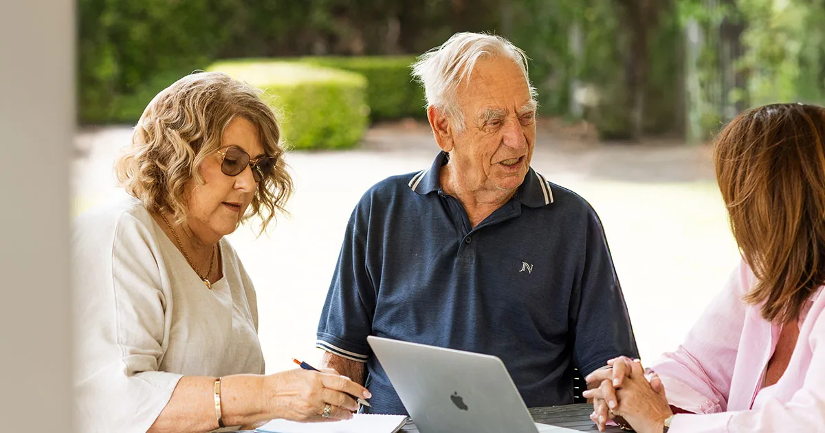 An older couple sit outdoors at a table with a laptop and notebook, discussing aged care fees with an advisor.
