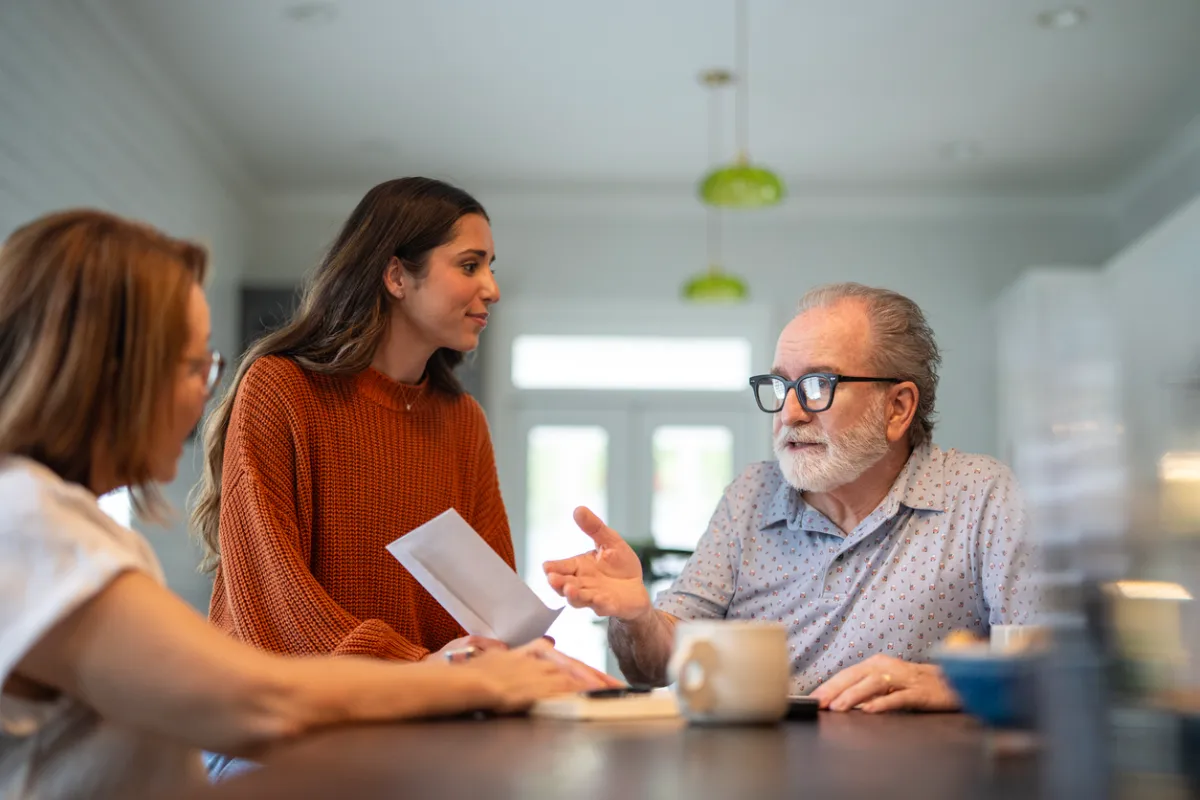 An older man and his daughter sitting together at the kitchen table, talking with an aged care assessor