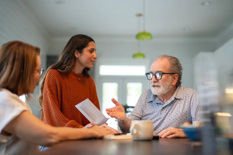 An older man and his daughter sitting together at the kitchen table, talking with an aged care assessor